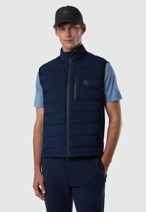 Uomo con berretto nero, camicia a maniche corte azzurro chiaro e gilet trapuntato blu navy con chiusura a zip e tasca sul petto, su sfondo grigio.