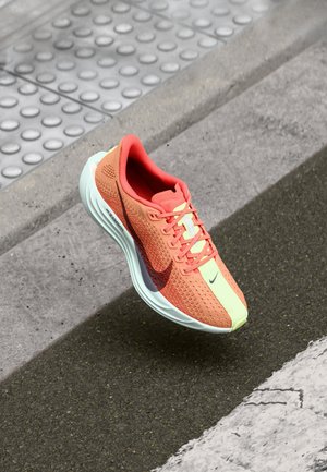 Koralowo i jasnozielony but biegowy Nike unoszący się nad szarym asfaltem obok krawężnika z powierzchnią dotykową i białym oznakowaniem drogowym.