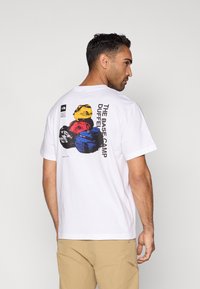 Λευκό βαμβακερό t-shirt με γραφική εκτύπωση πολύχρωμων σακιδίων πλάτης στην πίσω πλευρά, με το κείμενο «THE BASE CAMP DUFFEL» σε μαύρο.