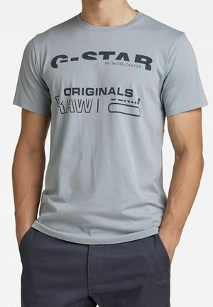 Homme portant un t-shirt gris clair de la marque G-STAR RAW avec un texte noir et un pantalon bleu marine, mains dans les poches, sur fond blanc.