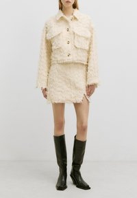 Crème-kleurige cropped jacket met twee voorzakken, gecombineerd met een mini rok. Beide zijn gemaakt van een textuur met franjes. Kniehoge zwarte laarzen.