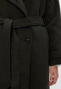 Casaco de lã verde escuro com cintura cinguada, grande colarinho e detalhes em botão. O tecido parece suave e texturizado.