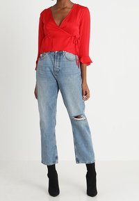 Haut en tissu rouge à manches longues avec des poignets à volants, associé à un jean bleu clair déchiré et taille haute. Bottines noires complètent la tenue.