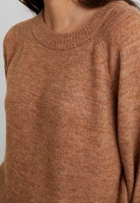 Pull en maille marron avec un col ras du cou côtelé, présentant un tissu doux et texturé ainsi qu'une subtile variation de couleur partout. Détail en gros plan.