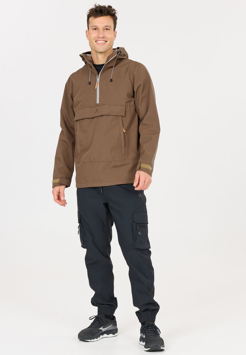 Hymyilevä mies seisoo, pukeutuneena ruskeaan huppariin, jossa on etutasku, ja mustiin kuorihousuihin, joissa on vetoketjutaskut, sekä mustat tennarit.