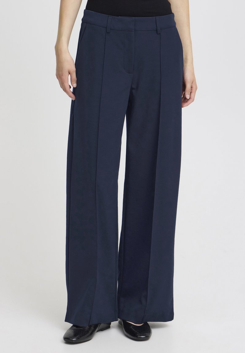 Femeie purtând pantaloni bleumarin cu talie înaltă, largi, cu pliuri frontale și pantofi negri ascuțiți, stând în picioare în fața unui fundal simplu.