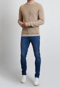 Pull en beige texturé avec un col rond et une bordure blanche, associé à un jean slim bleu et des baskets blanches.