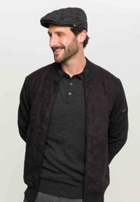 Giacca bomber nera trapuntata con colletto a coste sopra una polo scura. Un berretto piatto completa l'outfit, mostrando uno stile elegante e casual.