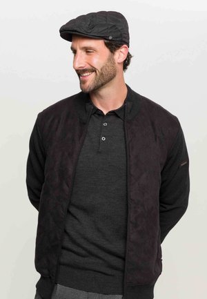 Jachetă bomber neagră matlasată cu guler ribbed peste un tricou polo închis. O pălărie plată completează ținuta, evidențiind un stil elegant și casual.