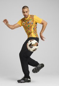 Multifarvet skjorte med gule og røde mønstre, sorte sweatpants, sorte fodboldstøvler, spiller der jonglerer med en hvid fodbold med grafiske detaljer.