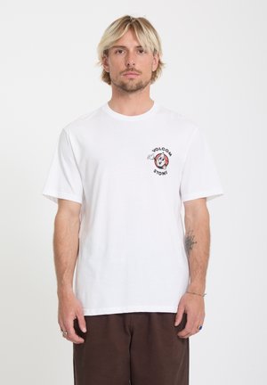Homme blond portant un t-shirt blanc Volcom Stone avec un logo cartoon sur la poitrine et un pantalon marron, debout devant un fond blanc uni.