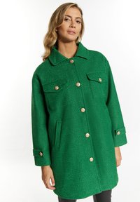 Cappotto verde testurizzato con colletto, due tasche sul petto e bottoni dorati. Maniche lunghe con polsini abbottonati.