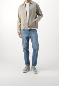 Beżowa lekka kurtka z zamkiem błyskawicznym, dwie boczne kieszenie, noszona na białej koszuli, zestawiona z jasnoniebieskimi dżinsami i szarymi tenisówkami.