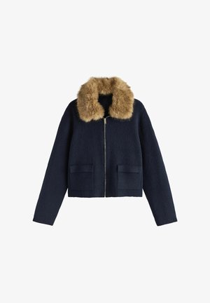 Mornarsko plava vunena skraćena jakna s ovratnikom od umjetnog krzna, prednjim zatvaračem na patentni zatvarač i dva teksturirana džepa s flasterima. Dugih rukava i kroja koji prati tijelo.