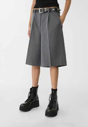 Personne portant un short large gris mi-cuisses, une ceinture noire cloutée, des bottes noires à plateforme lacées, et un haut noir avec les mains dans les poches.