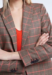 Bruine houndstooth blazer met rode en zwarte ruitpatronen; heeft gesneden revers en knoopmanchetten; gelaagd over een felrode top.