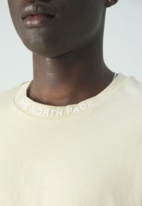 Světle béžové bavlněné tričko s žebrovaným výstřihem a logem "THE NORTH FACE" v bílé barvě. Hladký povrch, neformální design a pohodlný střih.