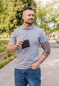 Black leather wallet held in hand, textured finish. Gray polo shirt, short sleeves. Defined arm with intricate tattoo pattern. Outdoor setting.