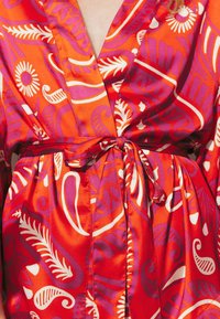 Robe en satin rouge ornée d'un motif floral coloré et abstrait en blanc et violet, avec une ceinture nouée à la taille et un décolleté en V.