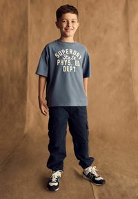 Modré bavlněné tričko s potiskem "SUPERDRY STATE PHYS. ED. DEPT", spárované s námořnickými cargo kalhotami a černobílými teniskami s akcenty.