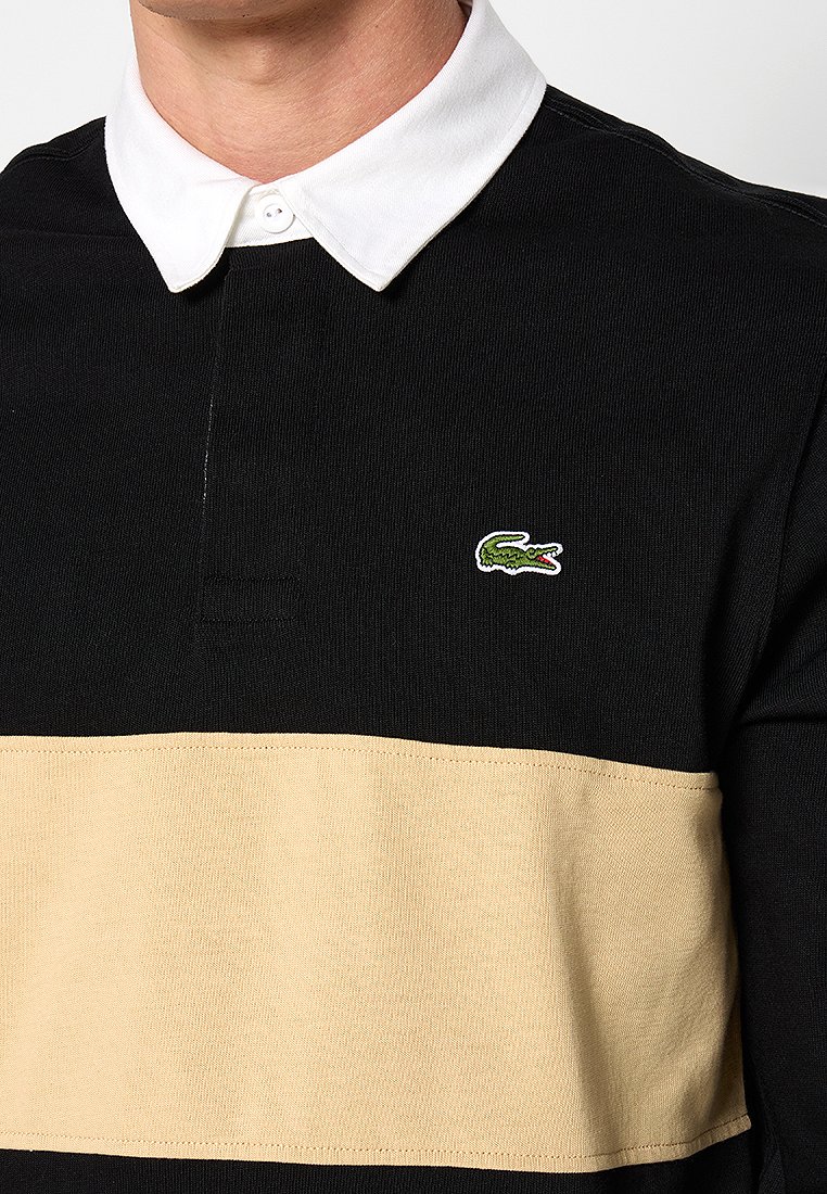 Polo à manches longues rayé noir et beige, avec un col blanc et un logo crocodile brodé vert sur la poitrine.