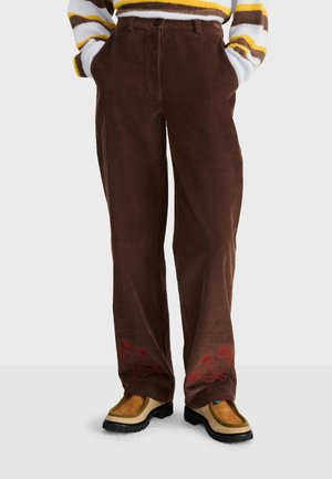 Pantalon en velours côtelé marron avec une broderie florale rouge près des ourlets, porté avec des chaussures beige et marron et un pull rayé partiellement visible.