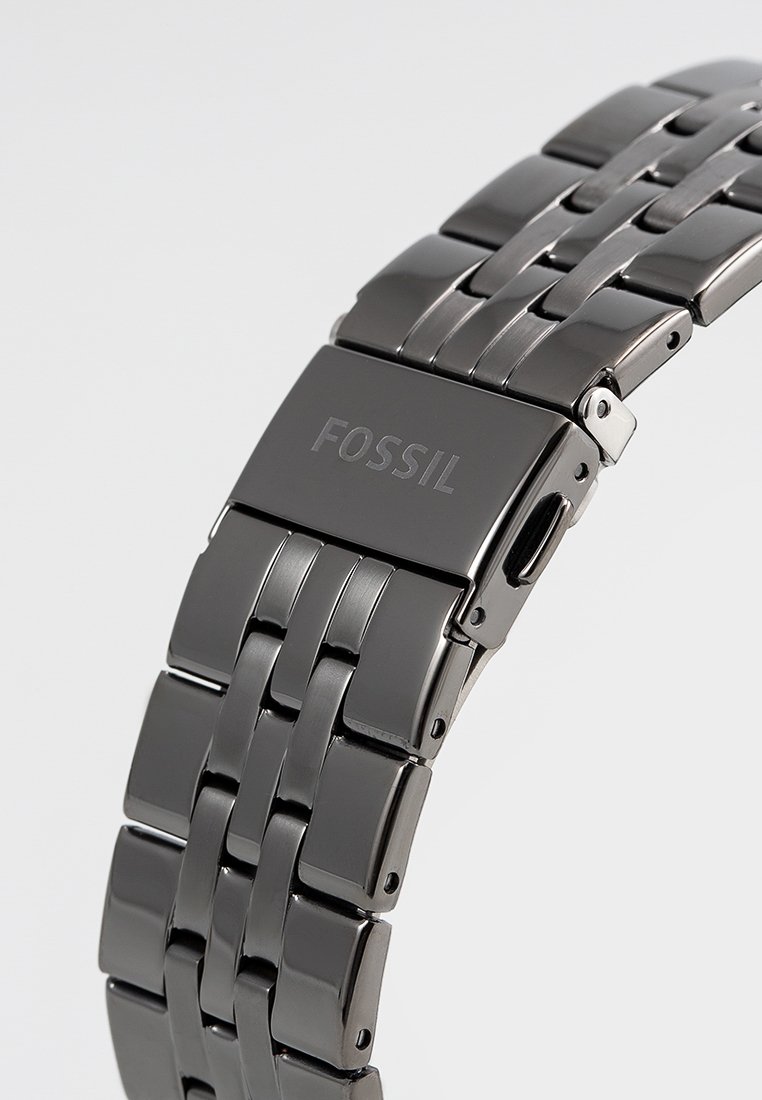 Banda de reloj de metal con un acabado negro pulido, que presenta un diseño de eslabones en forma de ladrillo y un cierre rectangular grabado con "FOSSIL."