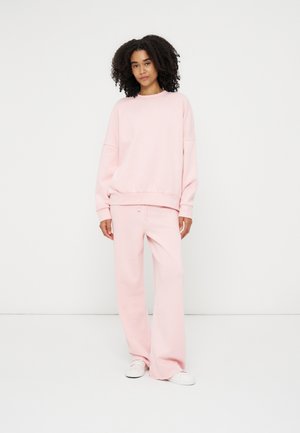 Kvinde med krøllet hår iført en løs lyserød sweatshirt og matchende brede bukser med hvide sneakers, stående mod en hvid baggrund.