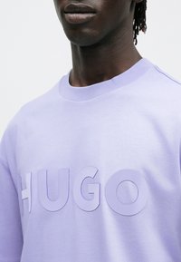 Μωβ βαμβακερό κοντομάνικο μπλουζάκι με μεγάλες, ανάγλυφες επιστημένες γράμματα "HUGO" σε ταιριαστή απόχρωση, με στρογγυλή λαιμόκοψη και απαλή υφή.