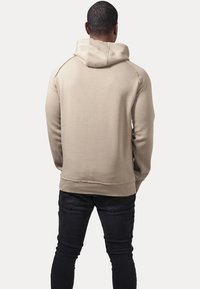 Beige hooded sweatshirt with a relaxed fit, featuring ribbed cuffs and hem, smooth texture, and flat seams, paired with dark jeans.