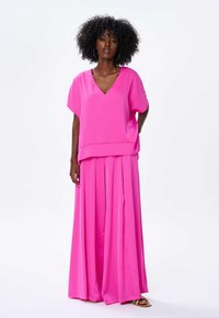 Jasnoróżowy, dwuczęściowy strój z bluzką z dekoltem w serek i krótkimi rękawami oraz spodniami z szerokimi nogawkami i plisami. Gładki materiał o miękkiej fakturze.