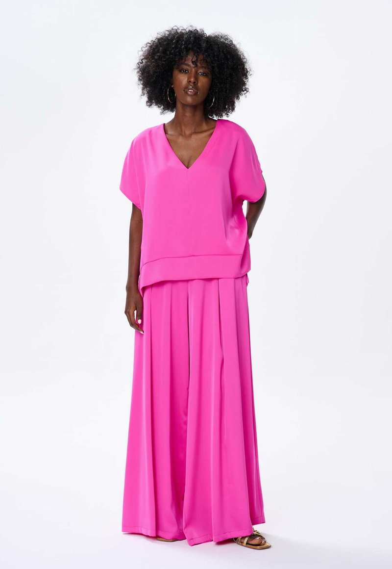 Jasnoróżowy, dwuczęściowy strój z bluzką z dekoltem w serek i krótkimi rękawami oraz spodniami z szerokimi nogawkami i plisami. Gładki materiał o miękkiej fakturze.