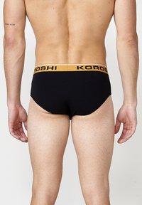 Schwarze Baumwollunterwäsche mit einem breiten gelben Bündchen, das das "KOROSHI"-Logo trägt. Die Rückseite zeigt einen sauberen, figurschmeichelnden Schnitt und eine glatte Textur.
