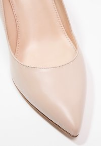 Escarpin en cuir beige à talon haut, avec un bout pointu, une texture lisse et un détail minimal de couture le long du bord.