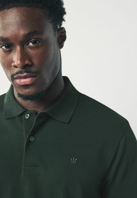 Dunkelgrünes Poloshirt aus strukturiertem Stoff, mit Kragen, zwei Knöpfen und einem dezenten goldenen Kronenlogo auf der linken Brust.