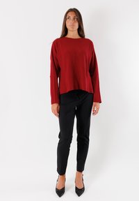 Donna in piedi contro uno sfondo bianco, che indossa un maglione rosso largo, pantaloni neri aderenti e tacchi a punta neri.