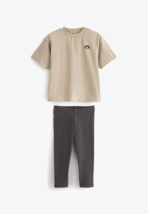 Bež majica kratkih rukava s okruglim izrezom i logotipom majmuna; uparena s tamno sivim tajicama koje imaju rebrastu teksturu.