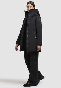 Frau in einer schwarzen Winterjacke mit Kapuze und Reißverschluss, die mit einem Fuß nach vorne auf einem weißen Hintergrund steht.