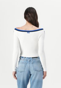 Hvit ribbet off-shoulder topp med blå og svart trimdetaljer, paret med blå høyt liv jeans, vist fra baksiden.