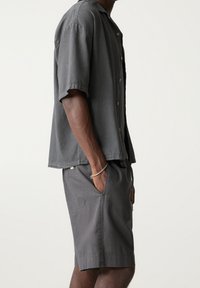 Camisa gris de manga corta con botones y corte relajado, combinada con pantalones cortos grises a juego. Ambas prendas están hechas de una tela ligera y texturizada.