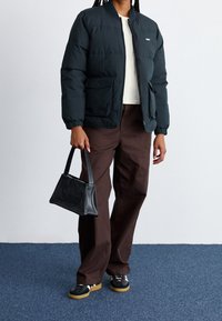 Giacca a vento verde scuro con polsini elasticizzati, due tasche anteriori. Pantaloni a gamba larga marroni. Borsa nera con logo in rilievo. Sneakers nere.