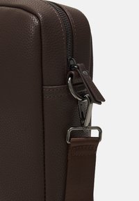 Coin de sac en cuir marron présentant une surface texturée, une fermeture éclair noire et un clip métallique attaché à une sangle marron avec une finition lisse.