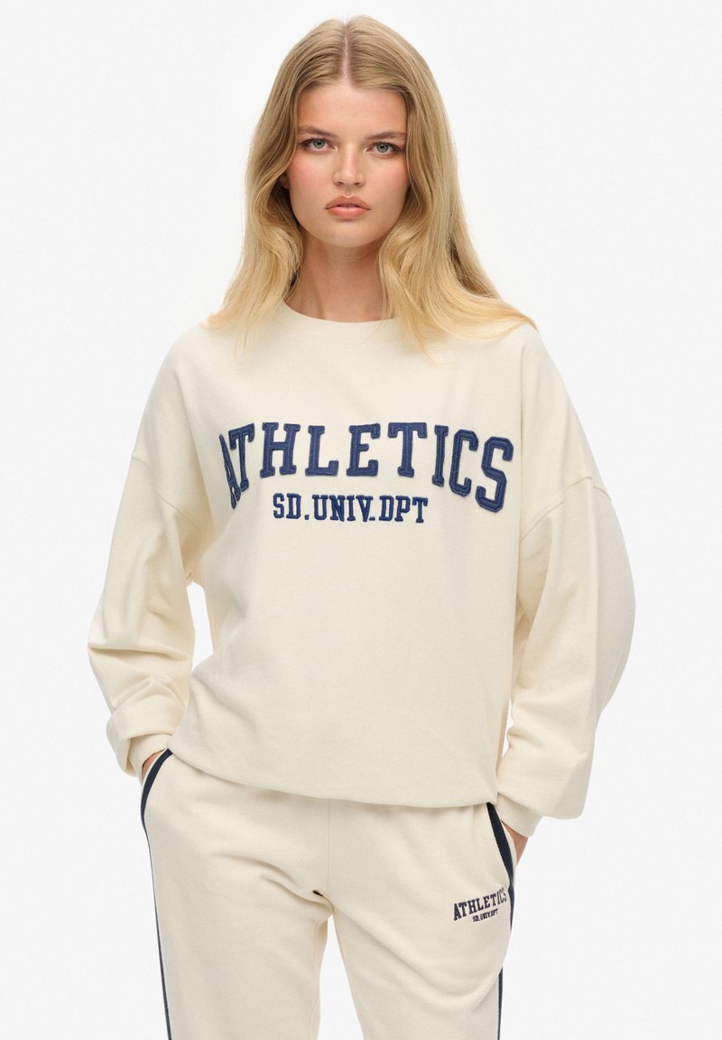 Sudadera crema con la palabra "ATHLETICS" en letras azul marino. Presenta mangas largas, un ajuste holgado y un diseño minimalista.