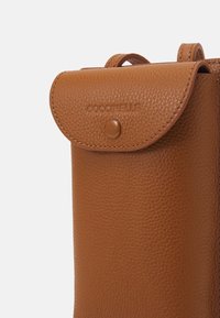 Brązowa skórzana saszetka o strukturalnej powierzchni, z zaokrągloną klapą i zapięciem na zatrzask. Wytłoczone logo marki z przodu.