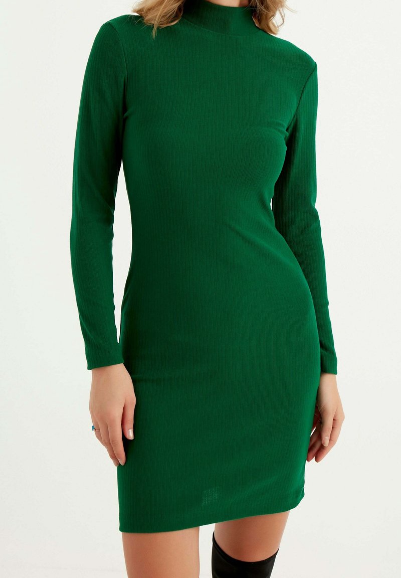 Femme portant une robe verte ajustée à manches longues et col montant, debout devant un fond blanc uni.