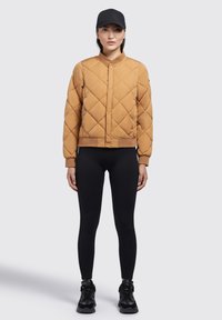 Gequilted, mosterdgele bomberjack met een geribbelde kraag en ritszakken, gecombineerd met zwarte leggings en sneakers.