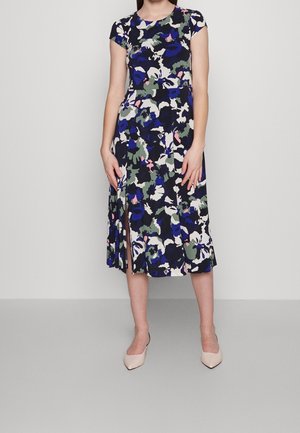 Mujer con vestido floral de manga corta hasta la rodilla con patrones en azul, verde, blanco y rosa, combinado con tacones beige de punta afilada.