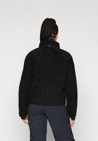 Černá kordurová bunda s vysokým límcem, žebrovanou strukturou a volným střihem, s knoflíkovým detailem na zadní straně.