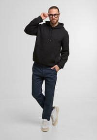 Felpa nera con cappuccio e motivo testurizzato, abbinata a pantaloni blu marine e sneakers bianche con accenti beige. Uomo in piedi in modo informale.