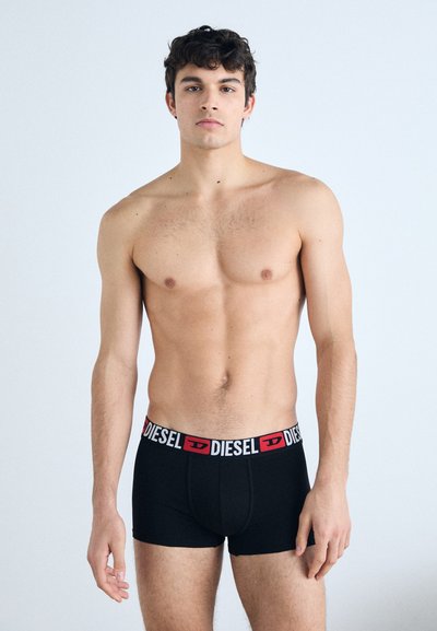 Boxer noirs avec une ceinture logo "DIESEL" rouge et blanche, présentant un design ajusté et une texture de tissu douce, modèle se tenant contre un fond uni.